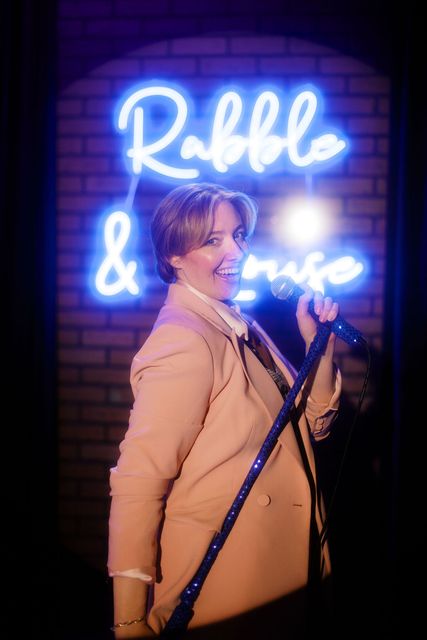 Katie at the mic in front of the neon Rabble and Rouse sign
